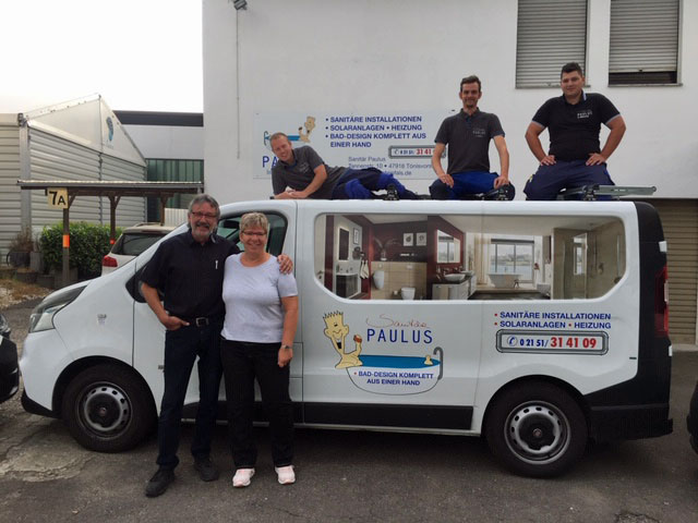 Teamfoto vor Firmenfahrzeug von Sanitär Paulus mit Bad-Design-Werbung, fünf Personen posieren lachend am und auf dem Auto.